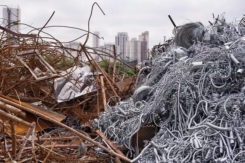 Large piles of mixed scrap metal, including rusted steel and shiny metal shavings, with city buildings in the background.