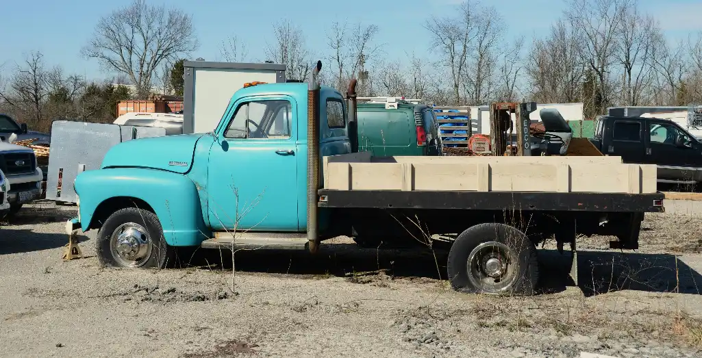 Vintage turquoise flatbed truck with a timber-sided tray parked in a gravel yard with other vehicles behind it